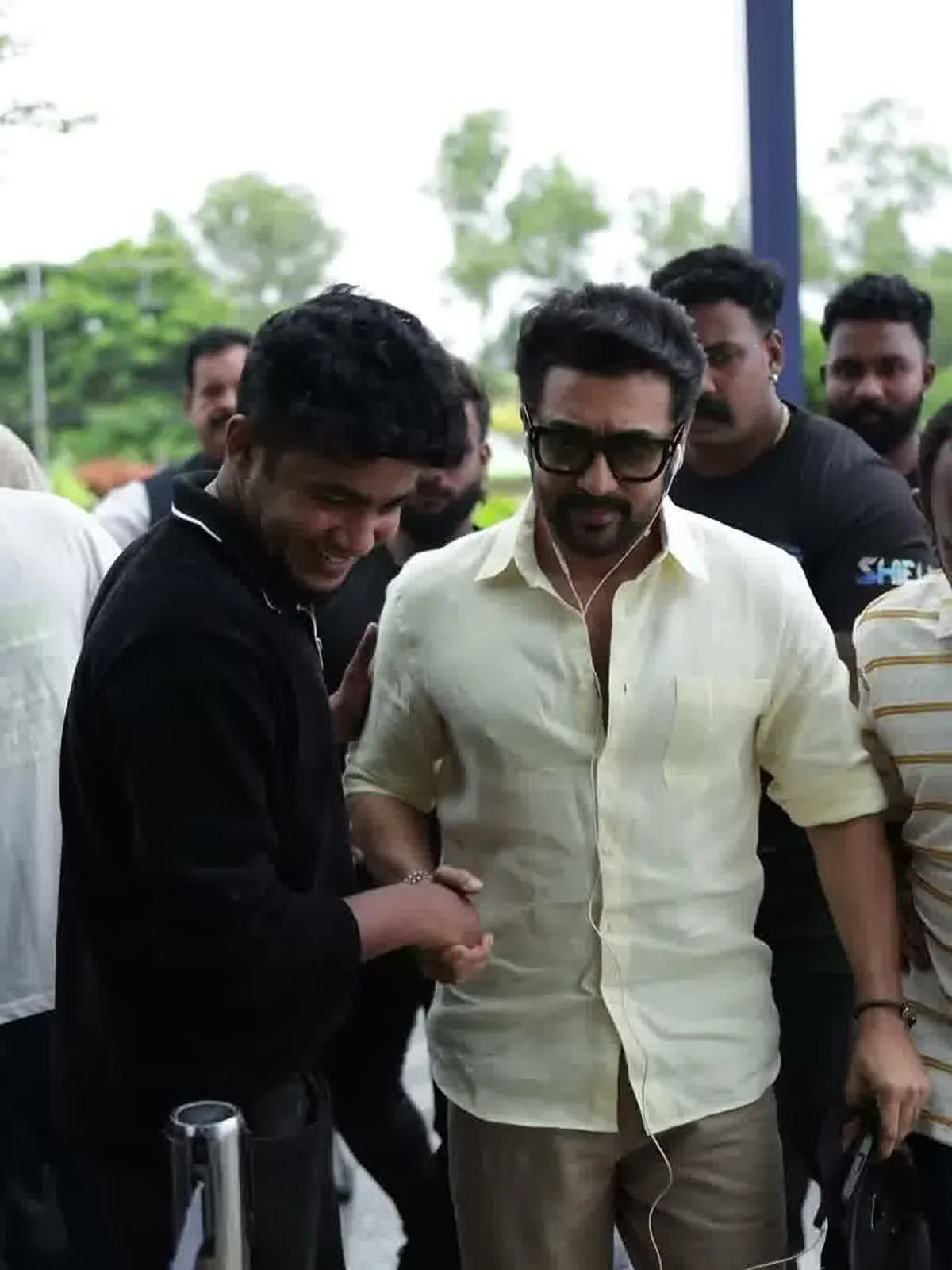 Suriya — Walking in — the sleeves rolled to just below the elbow, the shirt loose over the trousers